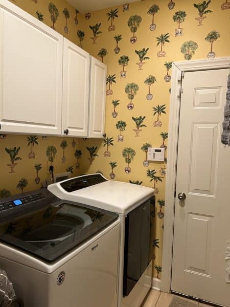 A laundry room featuring white cabinets, a washing machine, and a dryer. The walls are decorated with yellow wallpaper featuring palm tree designs.