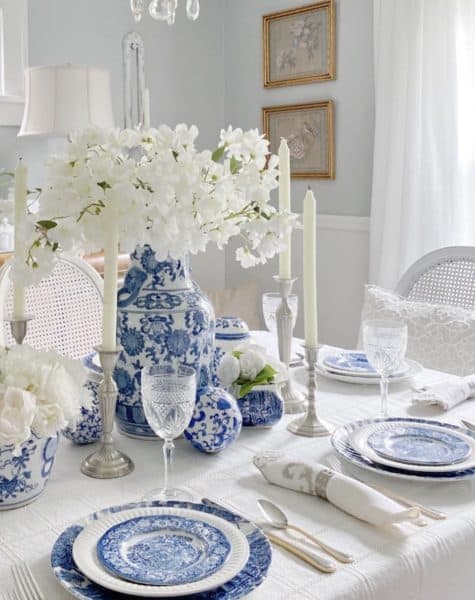 A beautifully set dining table featuring blue and white dinnerware, elegant glassware, and a centerpiece of white flowers in a blue vase, complemented by tall white candles.
