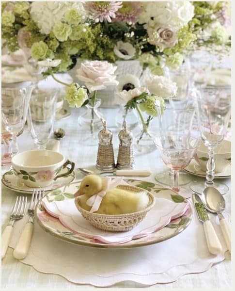 A beautifully set spring dining table featuring floral arrangements, elegant glassware, and a decorative spring-themed centerpiece with a small duck figurine.