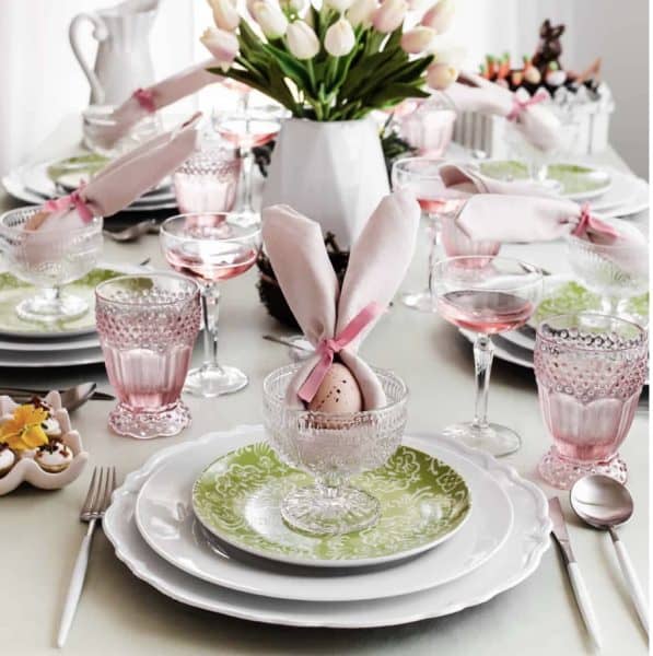 A beautifully set spring table featuring pastel-colored dinnerware, crystal glassware, and pink napkins resembling bunny ears, with a centerpiece of fresh tulips.