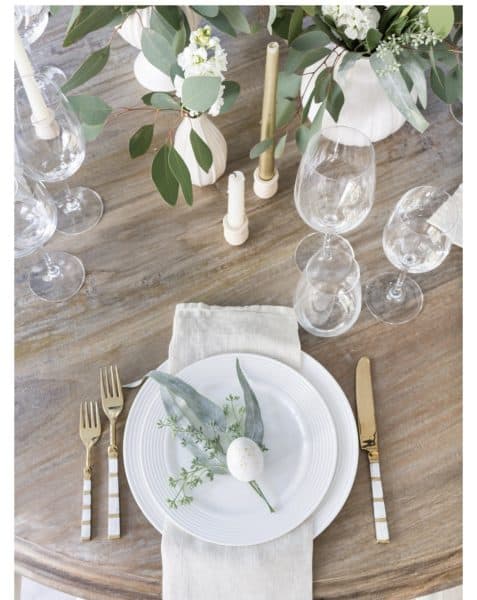 A beautifully arranged spring table setting featuring a white plate with a small decorative egg, surrounded by greenery and elegant gold cutlery, set on a wooden table with glassware and candles.