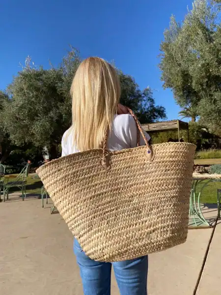 A woman with long blonde hair is standing outdoors, holding a large woven straw bag over her shoulder. Could be used as the basket for a Mother's Day Gift