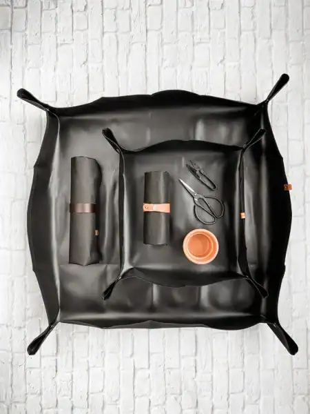 Flat lay of a gardening mat in black, featuring rolled gardening tools, a pair of scissors, and a small terracotta pot, set against a white brick background. perfect for a Mothers Day Gift Basket 