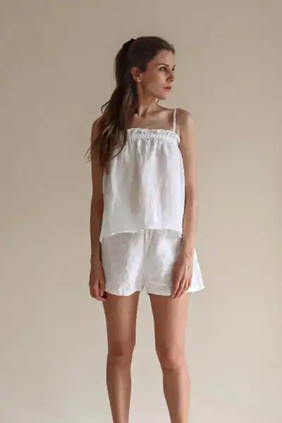 A woman wearing a white linen pajama set, consisting of a loose-fitting top and shorts, standing against a neutral background.