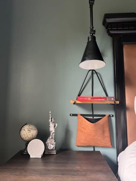 A nightstand in a boys bedroom, with a leather storage pouch for remotes to the TV and Fan, a shelf for books and a reading light