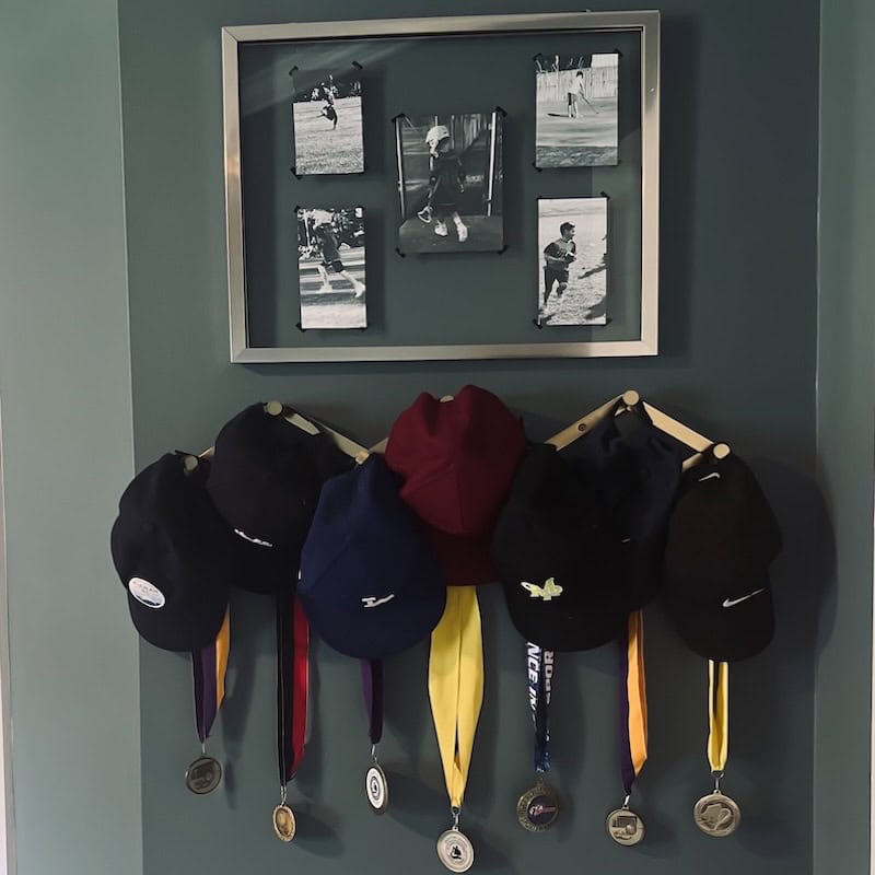 A collection of baseball or sports caps hanging on a wooden rack, accompanied by several medals, with a framed montage of black-and-white action photographs above.