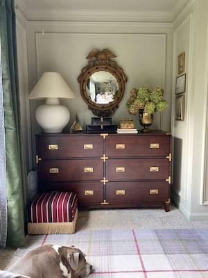 A stylish corner of a room featuring a wooden dresser with brass handles, a round mirror above it, a table lamp, and a vase of green flowers. A striped ottoman is beside the dresser, with a sleeping dog on a checkered rug.