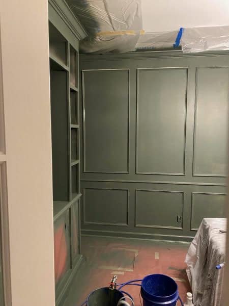 A partially finished room featuring green paneled walls and a built-in shelf next to an open space, with a painter's tarp on the floor and a blue paint bucket in the foreground.
