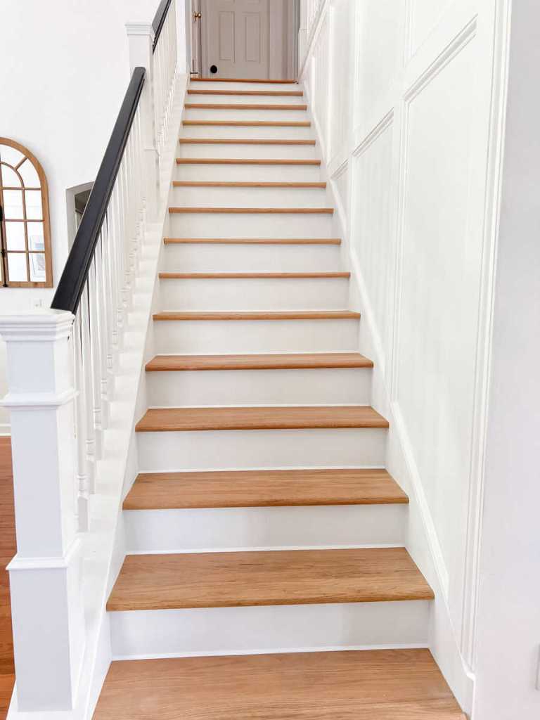 A well-lit stairway with wooden steps leading to an upper floor, featuring white walls and a black railing.