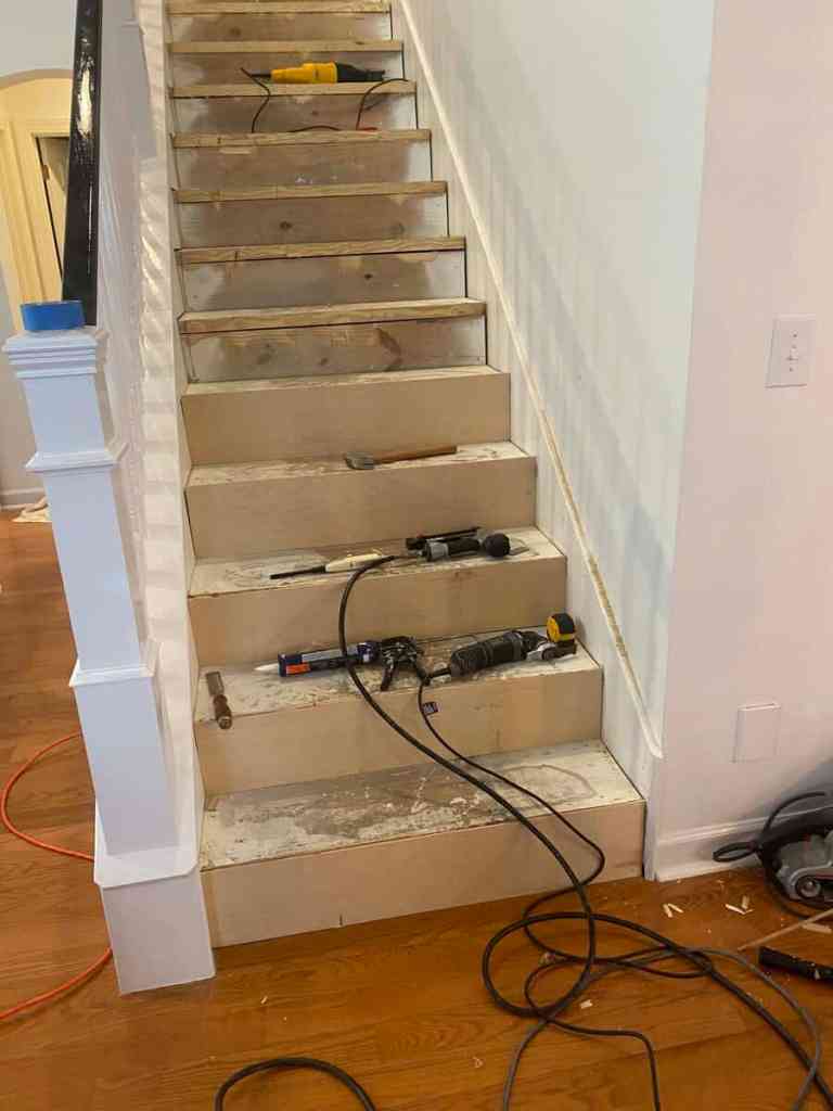 A partially renovated staircase with exposed wooden steps, tools including drills and a measuring tape scattered on the stairs, and wires visible on the floor.