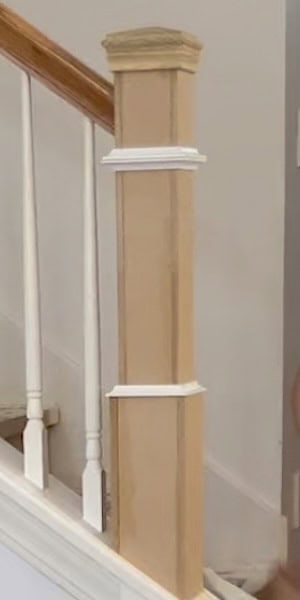 Close-up of a wooden stair railing post with white trim accents, situated beside a staircase.