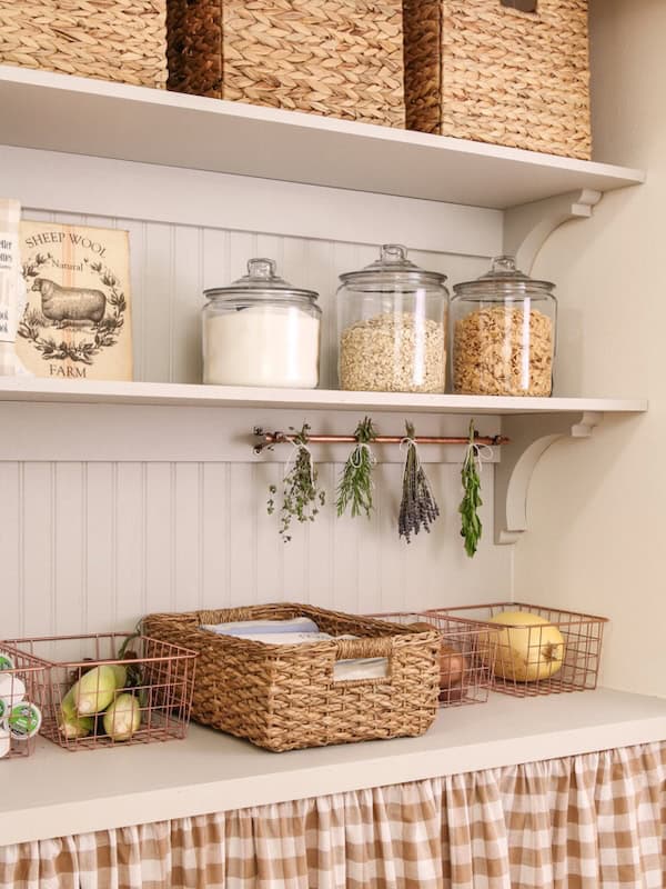 A kitchen shelf displaying glass jars filled with sugar, oats, and granola, alongside dried herbs hanging on a copper rod. Below, there are woven baskets containing fruits and papers, with a checkered fabric skirt covering the shelf.