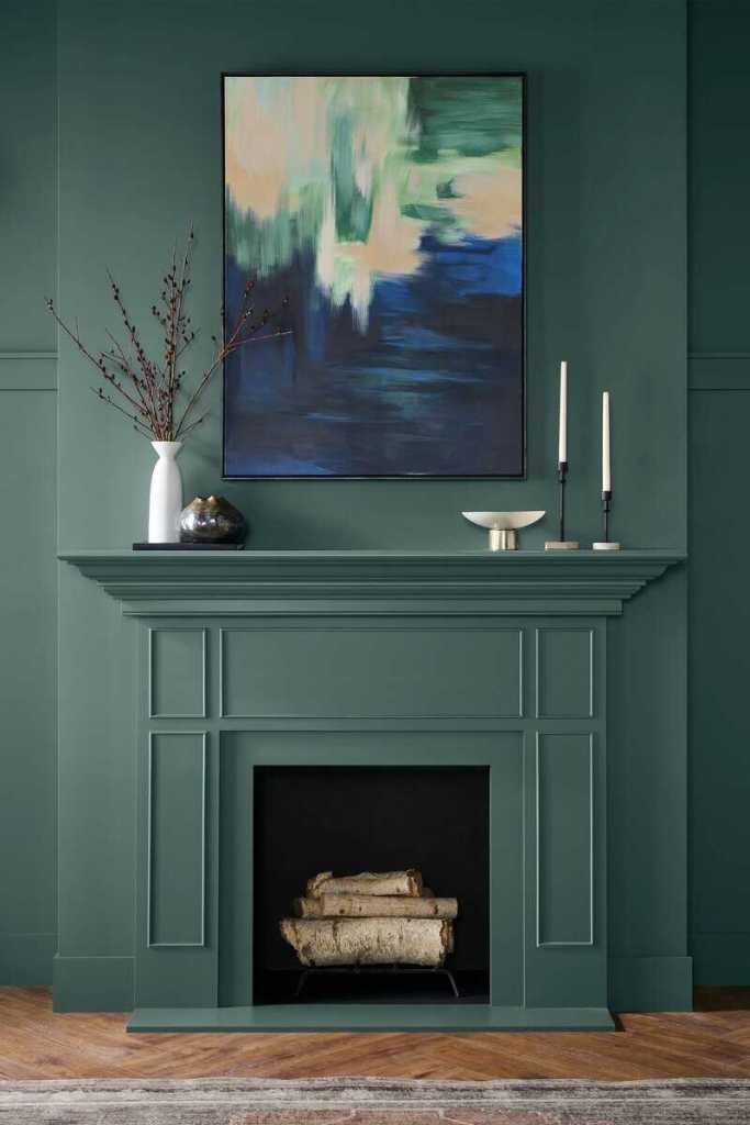 A modern fireplace mantel with a deep green wall, featuring an abstract painting above it, decorative items, and logs stacked in a black fireplace opening.