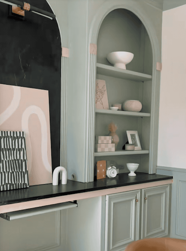 A stylish interior featuring a pale green cabinet with arched shelving, adorned with decorative items, including vases, art, and a small clock on a dark countertop.