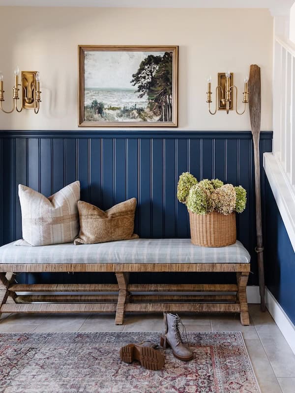 A cozy entryway featuring a wooden bench with cushions, a basket of flowers, framed artwork on the wall, and decorative sconces.