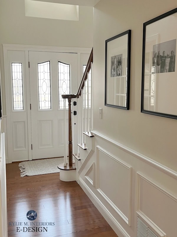 A well-lit entryway featuring a staircase with wooden railings, white walls, and decorative wainscoting. Large double doors with glass panels are visible, along with framed photographs on the wall and a small rug at the bottom of the stairs.