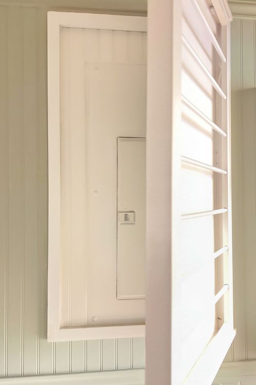 A partially open white wooden door or panel revealing a hidden compartment with a small latch, set against a light-colored wall with beadboard paneling.