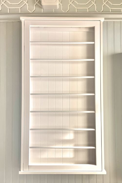 A white wooden display cabinet with multiple horizontal shelves, set against a light-colored wall with a decorative pattern.