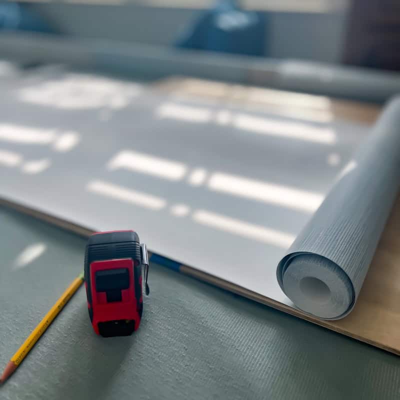 A red and black tape measure next to a yellow pencil on a textured surface, with a roll of gray and white paper in the background and sunlight casting shadows.