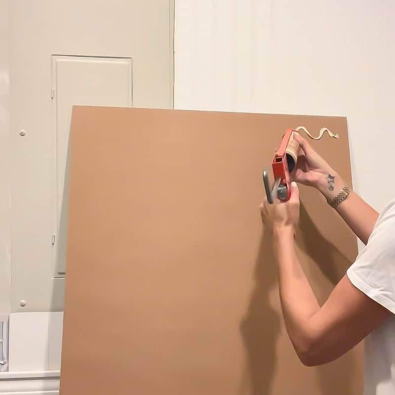 A person using a tape dispenser to apply tape on a large brown paper board mounted against a wall.