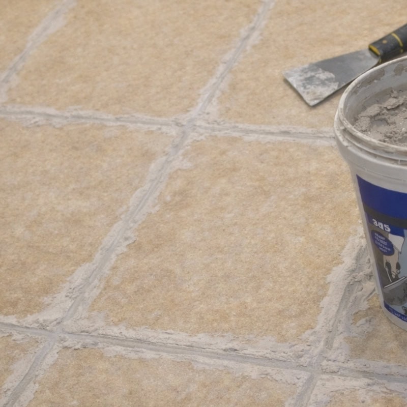 Close-up of a tiled floor with light-colored grout lines, a plastic bucket of material, and a trowel resting on the floor.