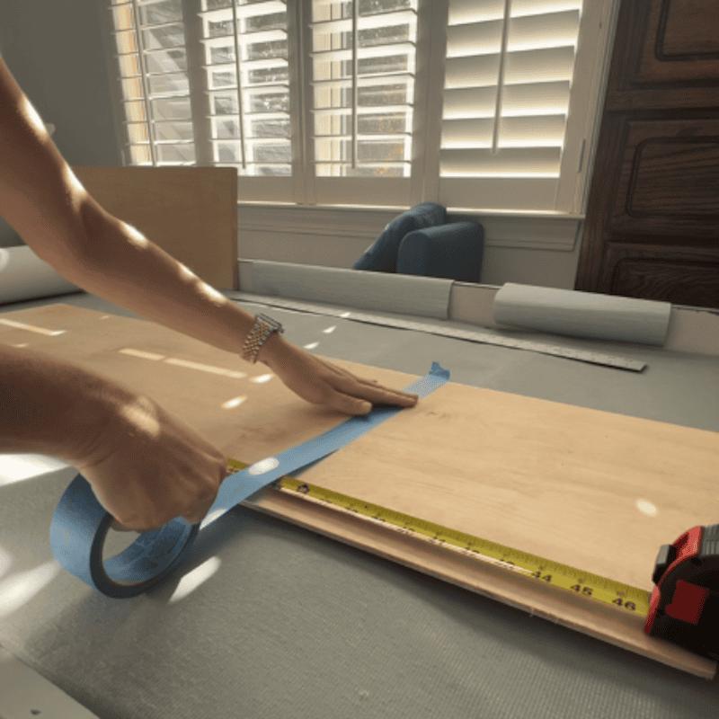 A person applying blue painter's tape to a wooden board while measuring with a tape measure on a work surface.