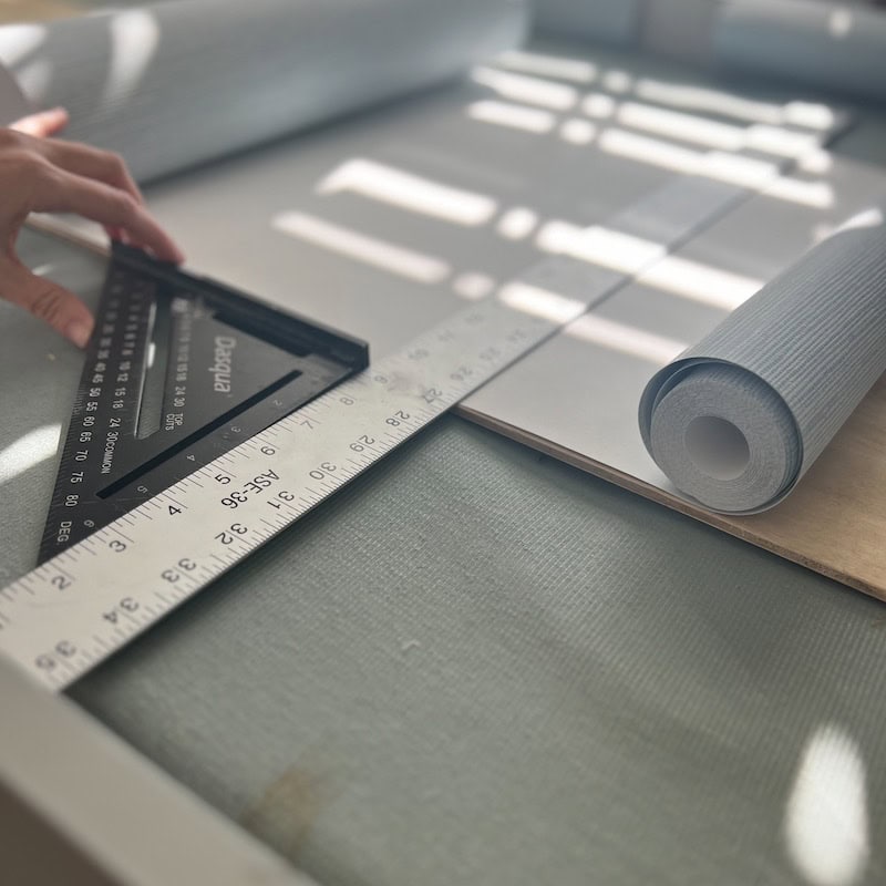 A close-up of a hand holding a triangle ruler next to a measuring tape, cutting a roll of wallpaper