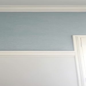 Blue grasscloth wallpaper in a corner of a white wall, with trim installed 3/4 up the wall, ready for paint to be applied. This image showcases the process of wallpaper and matching paint for a cohesive, stylish room design.