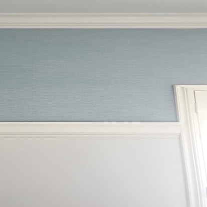 Blue grasscloth wallpaper in a corner of a white wall, with trim installed 3/4 up the wall, ready for paint to be applied. This image showcases the process of wallpaper and matching paint for a cohesive, stylish room design.