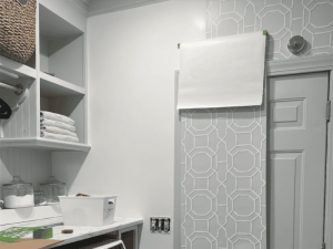 A laundry room featuring white shelving with neatly stacked towels, a transparent storage container, and a blank sheet of paper taped to the wall beside a door with decorative wallpaper.