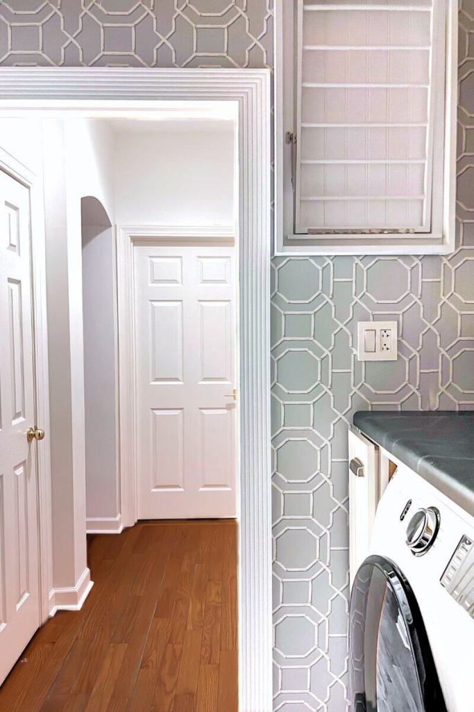 A modern laundry room with patterned gray wallpaper, featuring a white washing machine, wooden flooring, and a doorway leading to a hallway.