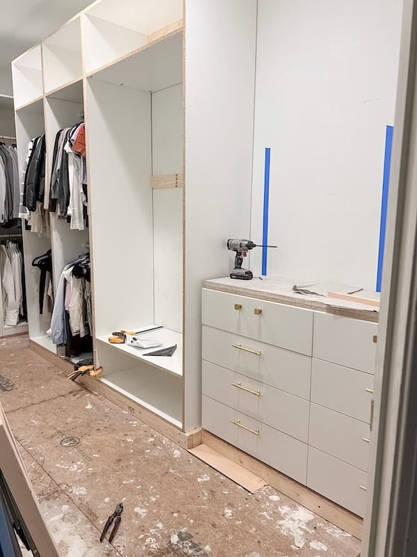 A partially completed interior space featuring a closet with hanging clothes on the left and a set of white drawers on the right. The floor is unfinished, and tools are visible on the counter and ground.