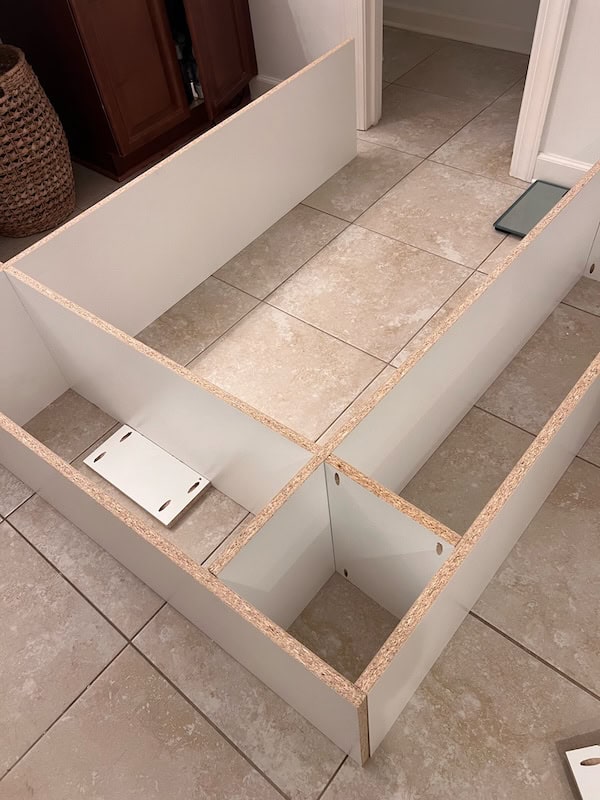 A partially assembled white furniture frame on tiled flooring, featuring several open sections and a connector piece in the center.