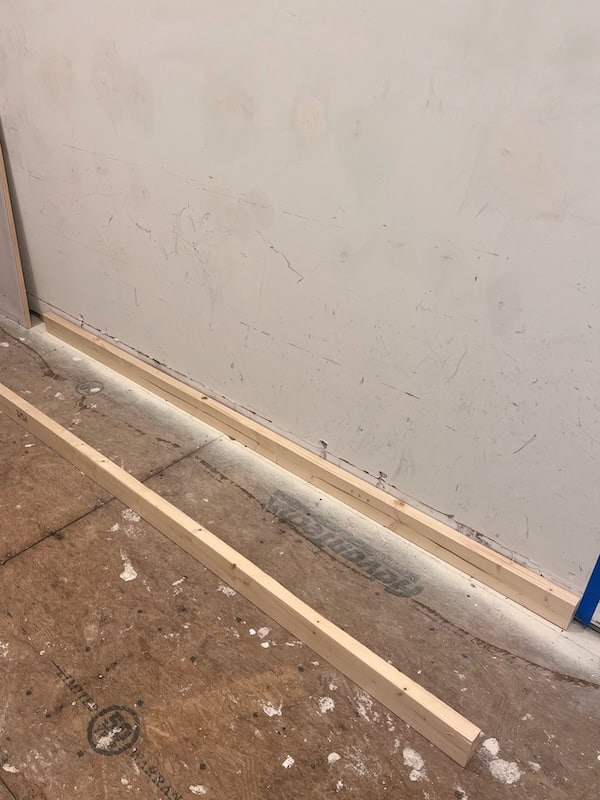 A wooden board lying on a construction floor next to a wall with visible marks and scratches.