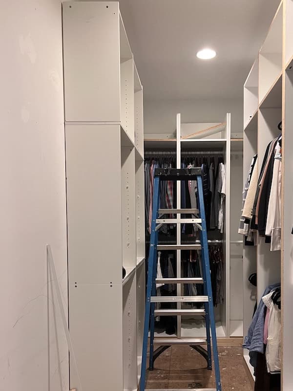 A partially organized closet featuring new white shelving units, a blue step ladder in the center, and hanging clothes on the right side.