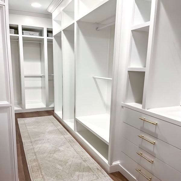 A spacious, modern walk-in closet with white walls and shelving, featuring a central rug and drawers with gold handles.