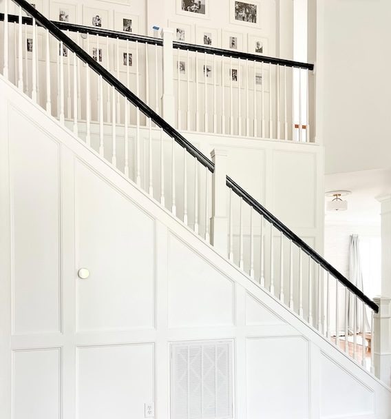 Interior view of a white-painted staircase with black railing, featuring wainscoting on the walls and framed photographs in the background.