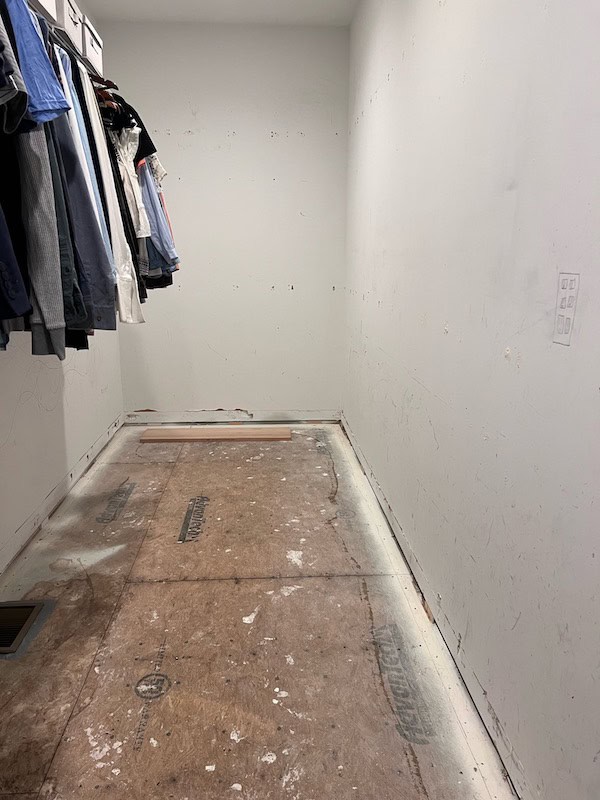 Interior of a partially finished closet with hanging clothes, bare walls, and a wooden plank on the floor.