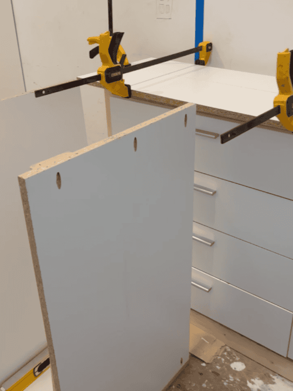 A close-up view of a partially assembled cabinet with a clamped top and a side panel leaning against it. The cabinet has white surfaces and is situated in a workshop environment.