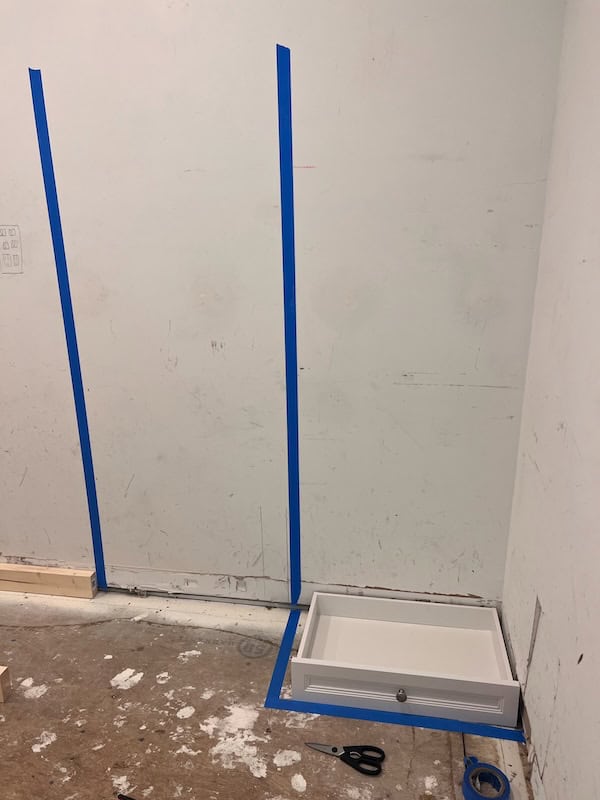 A room under renovation featuring blue painter's tape on the wall, a white drawer placed on the floor, and a pair of scissors nearby.