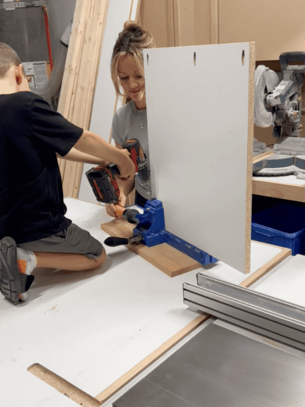 A child and an adult woman working together in a workshop, using a power drill on a piece of white board secured in a drilling jig, with woodworking tools and materials in the background.