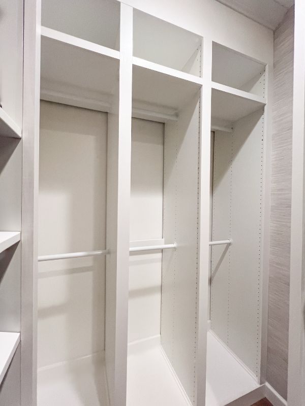 After photo of a  renovated narrow primary walk-in closet frames for hanging space, painted white with grasscloth wallpaper
