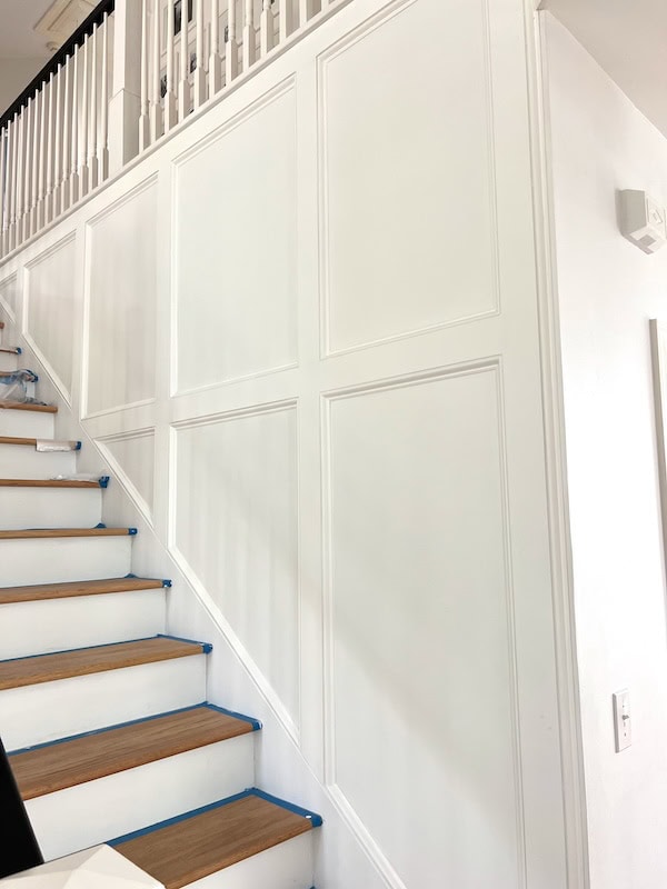 A staircase with wooden steps and white walls featuring decorative paneling, partially covered with painter's tape.