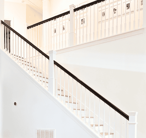 Interior view of a staircase with white railings and wooden steps, leading to an upper level, against a light-colored wall.