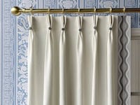 Close-up of cream-colored curtain panels with pleats and decorative trim, hanging on a gold curtain rod against a patterned blue wallpaper background.