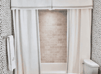 A well-designed bathroom featuring white curtains framing a bathtub, with a textured tiled back wall and a clean, modern toilet.
