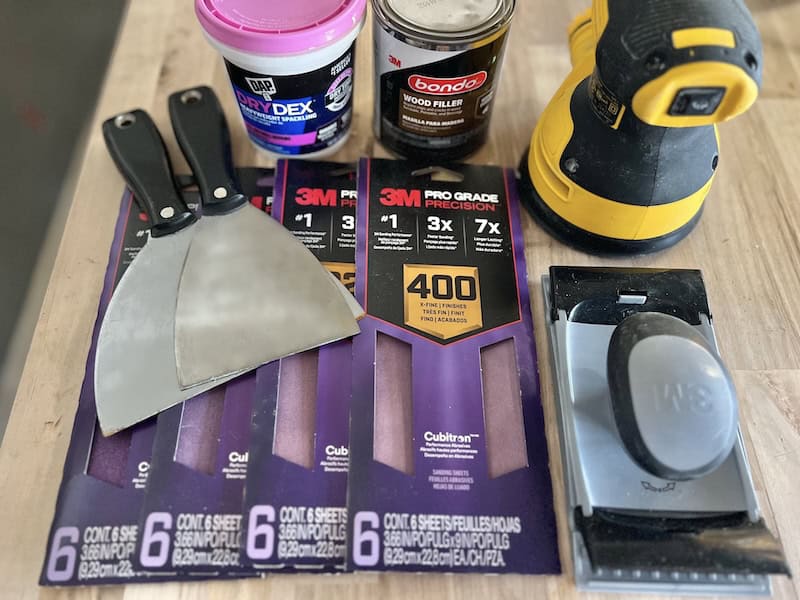 An assortment of painting and woodworking tools, including two putty knives, a can of wood filler, a container of spackling paste, sandpaper packs, and an electric sander, all arranged on a wooden surface.