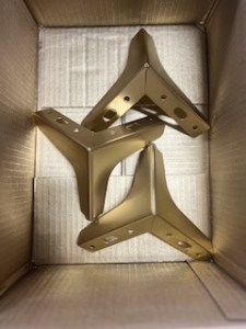 Three gold metal brackets arranged in a cardboard box.