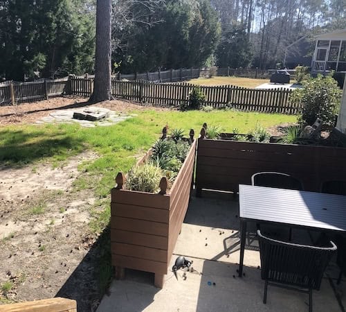 A backyard scene featuring a small patio with a table and chairs, surrounded by landscaping and a fire pit area in the background. The grass is lush, and there are wooden garden boxes with plants, indicating a cozy outdoor space.