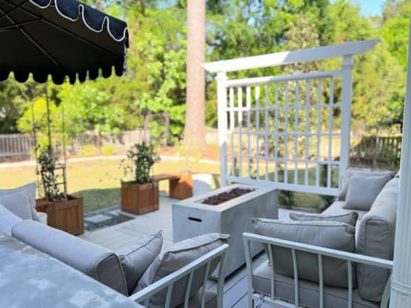 Cozy backyard seating area featuring a white patio set, an outdoor fire pit, potted plants, and a trellis in a lush green environment.